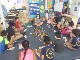 Teacher and students all with masks on, sitting on the floor during a learning exercise.