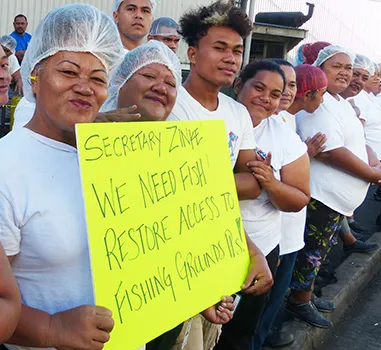 Cannery workers hold up sign reading "We need fish, restore access to fishing grounds, pls!"