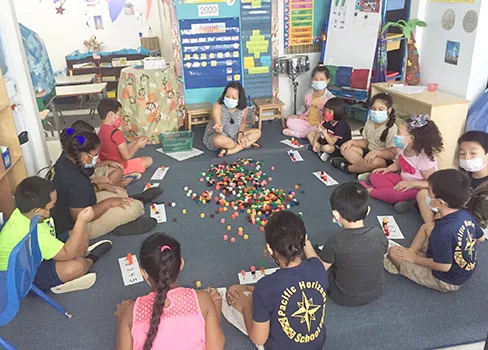 Teacher and students all with masks on, sitting on the floor during a learning exercise.