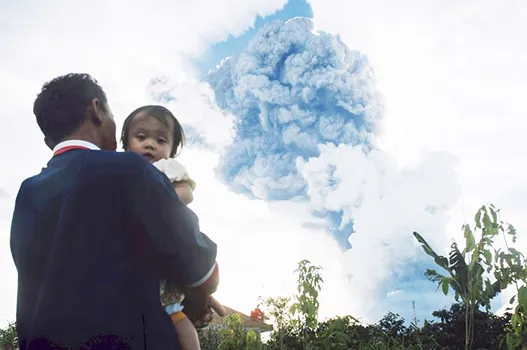 In this Sunday, June 9, 2019, file photo, a man carrying a child watches Mount Sinabung spew volcanic material into